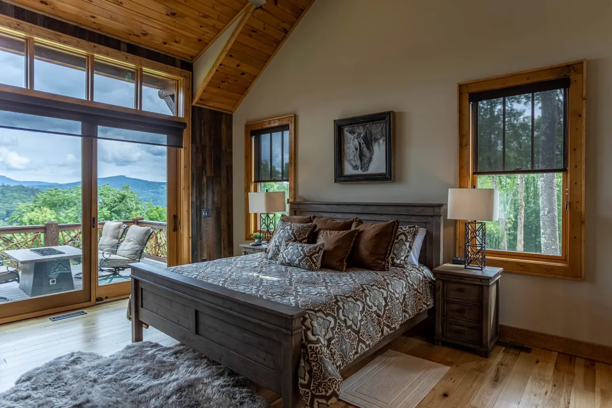 A king suite bedroom with a bed adorned in decorative pillows, large windows offering an inspiring view of the mountains, wooden furniture, and tasteful wall art. The soft natural light creates a serene, tranquil atmosphere, perfect for restful sleep and relaxation.