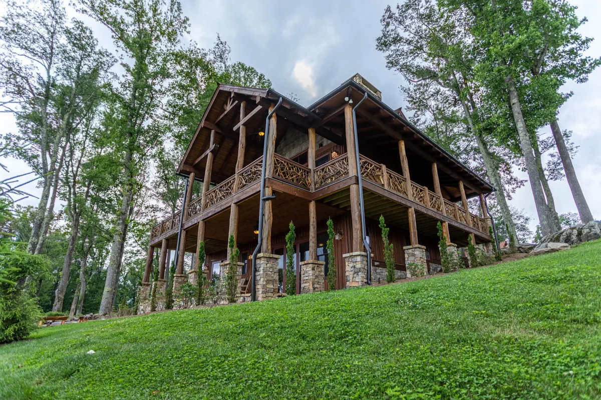 The image showcases the exterior of Lazy Bear Cabin, a beautiful wooden structure set atop sturdy stone pillars, surrounded by lush greenery. The inviting porch offers the perfect spot to relax and take in the sweeping mountain views, while the spacious yard hints at space for outdoor gatherings and sunset barbecues.
