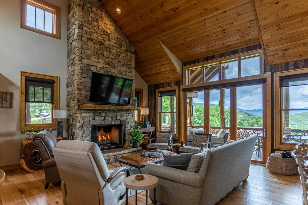 A warm, inviting second living room featuring a prominent stone fireplace, large windows letting in natural light, comfortable seating, and a television. The space exudes coziness and is clearly designed for relaxing evenings, movie nights, or gathering with family and friends.