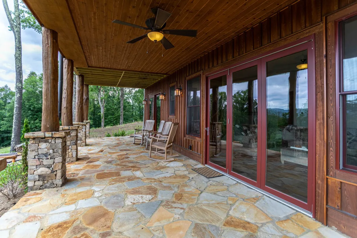 A tranquil porch scene featuring sturdy wooden pillars, a stone pathway, and classic rocking chairs facing outward. Large glass doors connect the porch to the interior, and mature trees frame the setting, suggesting immediate access to nature.