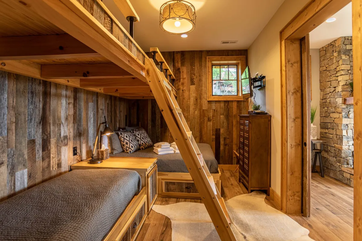 A cozy bunk room featuring a sturdy wooden bunk bed, rustic wood-paneled walls, a window letting in natural light, and a warm lighting fixture. The space feels inviting and perfect for children or groups to share laughter and nighttime chats.