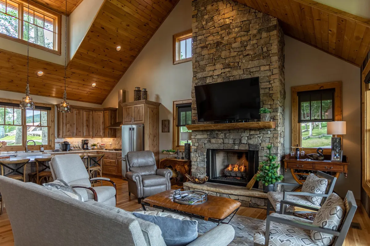 This image captures an inviting open-concept living space with a prominent stone fireplace, large windows flooding the area with natural light, a spacious comfortable seating area, and a wooden dining table that connects seamlessly to the kitchen. The setting exudes warmth and sociability, perfect for gatherings and relaxed evenings.