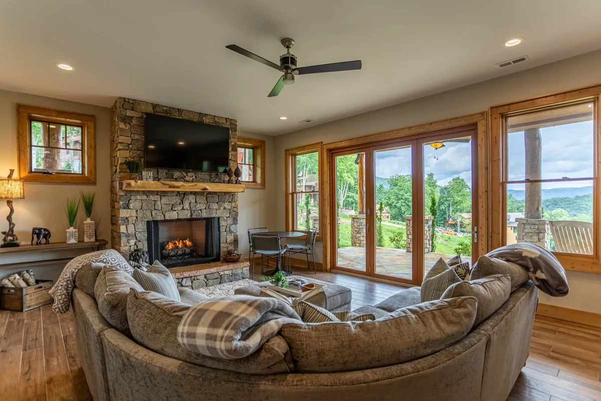 A spacious living area featuring a stone fireplace, large sectional sofa, floor-to-ceiling windows, and wooden accents. The space is ideal for socializing, relaxing after outdoor adventures, or hosting group activities indoors, with views of sunsets through the windows.