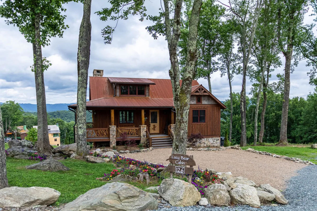 A charming cabin exterior with a welcoming wooden porch, flower beds, natural rock formations, and a signpost. The porch is set against a backdrop of greenery, inviting guests to step out and explore the surrounding trails and natural landscape.