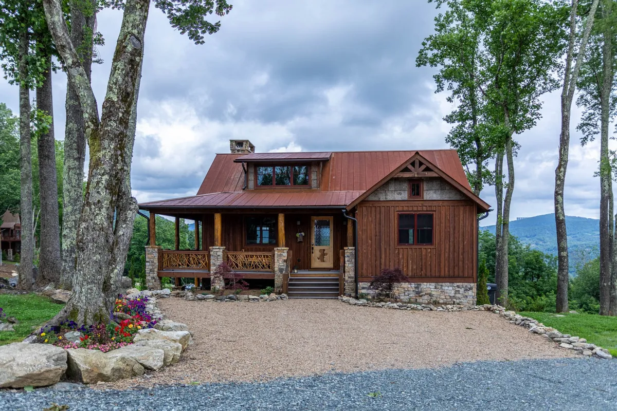 The cabin’s wooden façade, metal roof, stone porch, and surrounding trees are showcased, with a backdrop of mountain views. The inviting porch and garden set the scene for gatherings and tranquil moments outdoors.