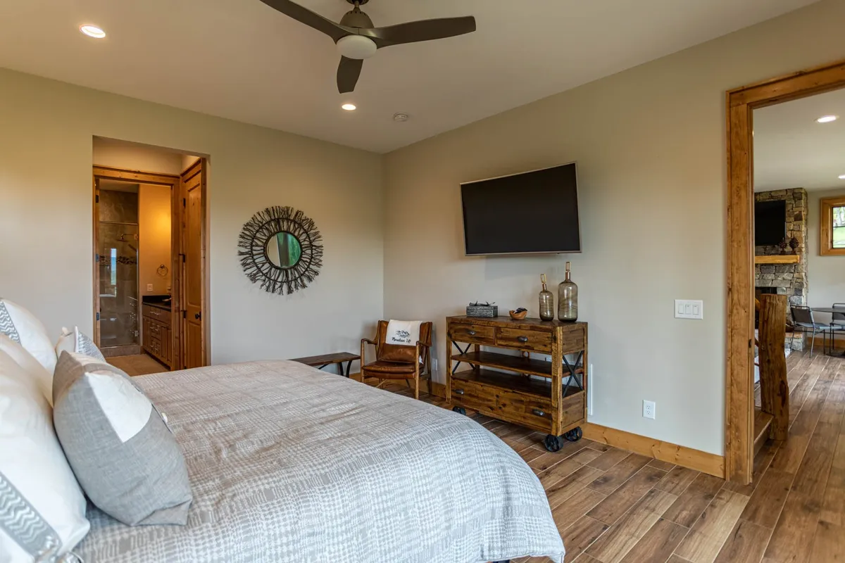 A king suite bedroom featuring patterned luxury bedding, a large wall-mounted TV, a spacious mirror, and wooden furniture. The space glows with natural light, offering a peaceful spot for unwinding with a show or a book.